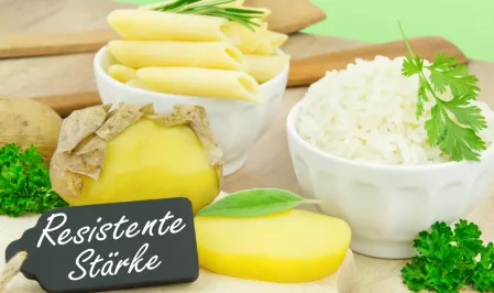 Resistente Stärke geschälte Kartoffeln auf einem Brett, Nudeln und Reis in zwei weißen Schalen. Davor steht ein Schild mit der Aufschrift Resistente Stärke